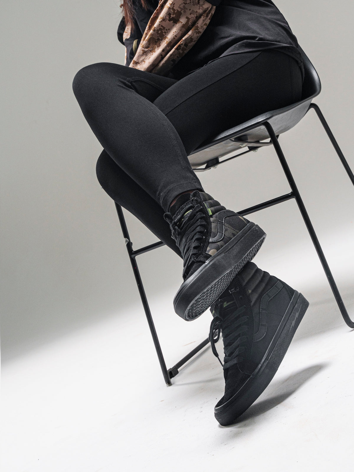 Model seated in a chair wearing black and camouflage DEFCON Syndicate SK8-Lo shoes, with the outsole visible.