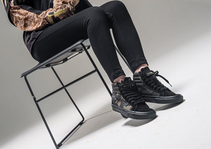 Model seated against a light studio backdrop wearing a pair of black and camouflage DEFCON Syndicate SK8-Lo shoes.