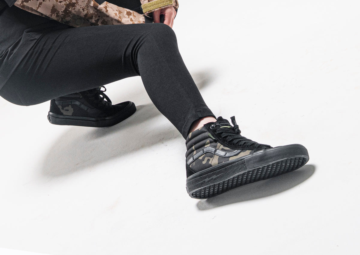 Model wearing black and camouflage DEFCON Syndicate SK8-Lo Notchback shoes, showing the side profile and treaded sole.