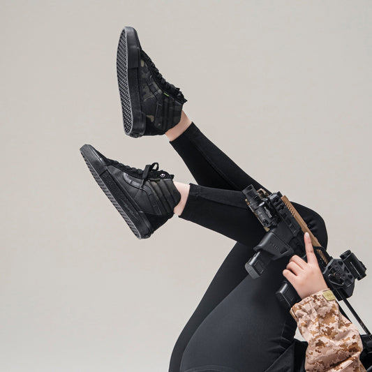 Model wearing black DEFCON Syndicate SK8-Lo Notchback shoes with black leggings, shown in a studio pose from below.