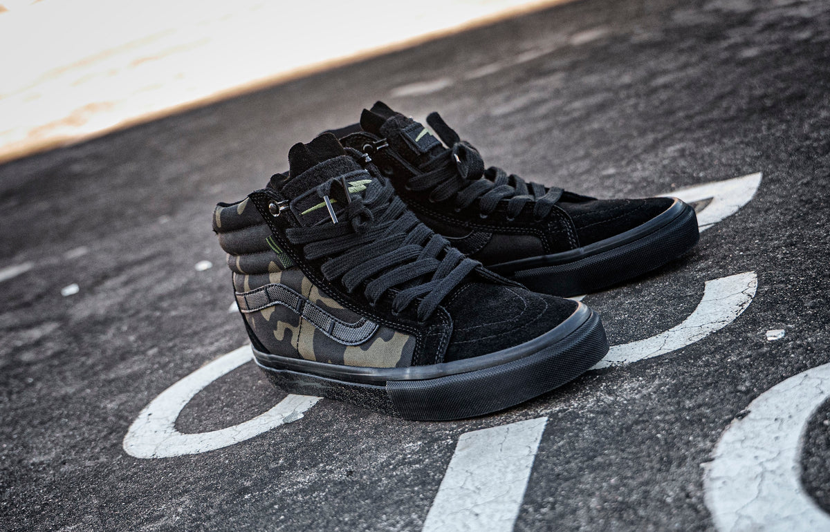 Pair of black and camouflage DEFCON Syndicate SK8-Lo Notchback shoes on pavement, showing the side panels and treaded soles.