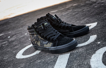 Pair of black and camouflage DEFCON Syndicate SK8-Lo Notchback shoes on pavement, showing the side panels and treaded soles.