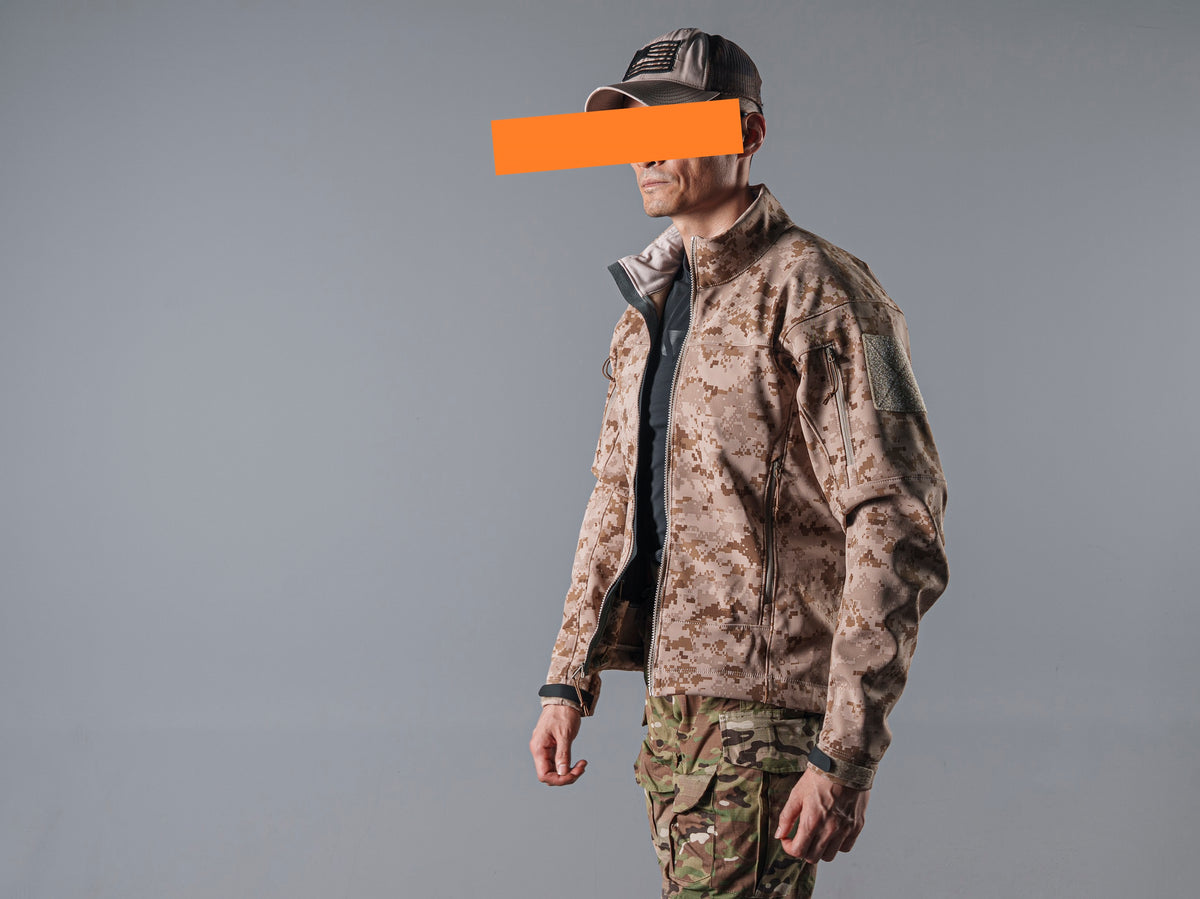 Rear three-quarter view of a model wearing the tan AOR1 digital camouflage Minotaur softshell jacket with a raised collar.