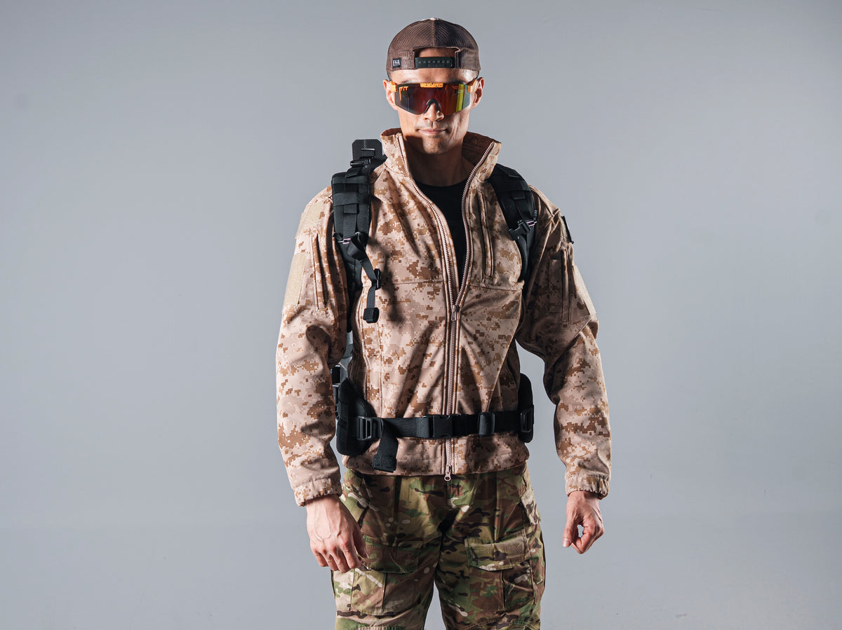 Front view of a model wearing the tan AOR1 Cold Fusion softshell jacket with black backpack straps and a waist belt.