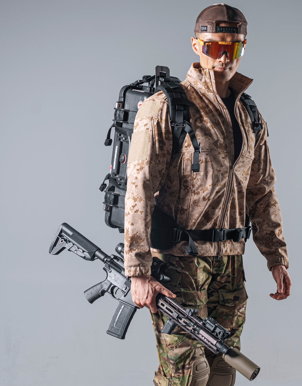 Three-quarter view of a model wearing the tan AOR1 Cold Fusion softshell jacket and carrying a rifle beside a black hard-shell pack.