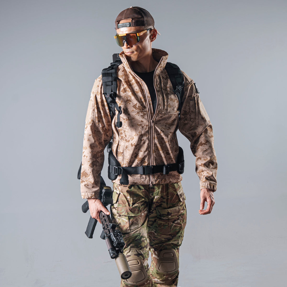 Model wearing the tan AOR1 digital camouflage BKG BE-Style Cold Fusion softshell jacket with a black tactical backpack and waist belt.