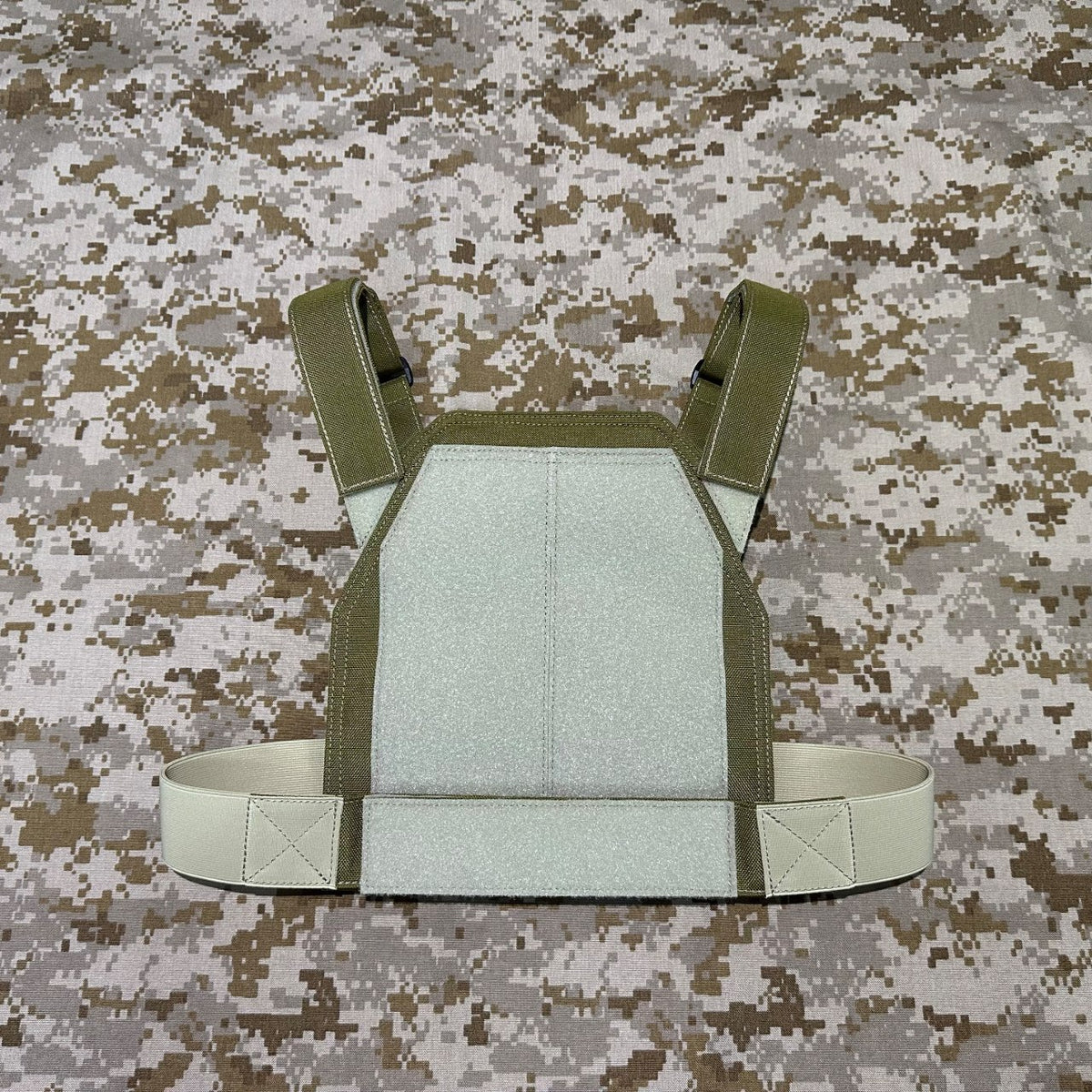 Front view of the olive low-vis plate carrier showing its pale rectangular panel and shoulder straps on a camouflage background.