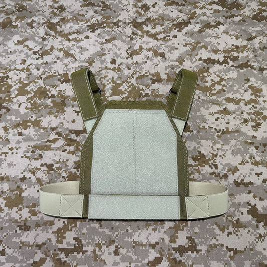 Front view of the olive low-vis plate carrier showing its pale rectangular panel and shoulder straps on a camouflage background.